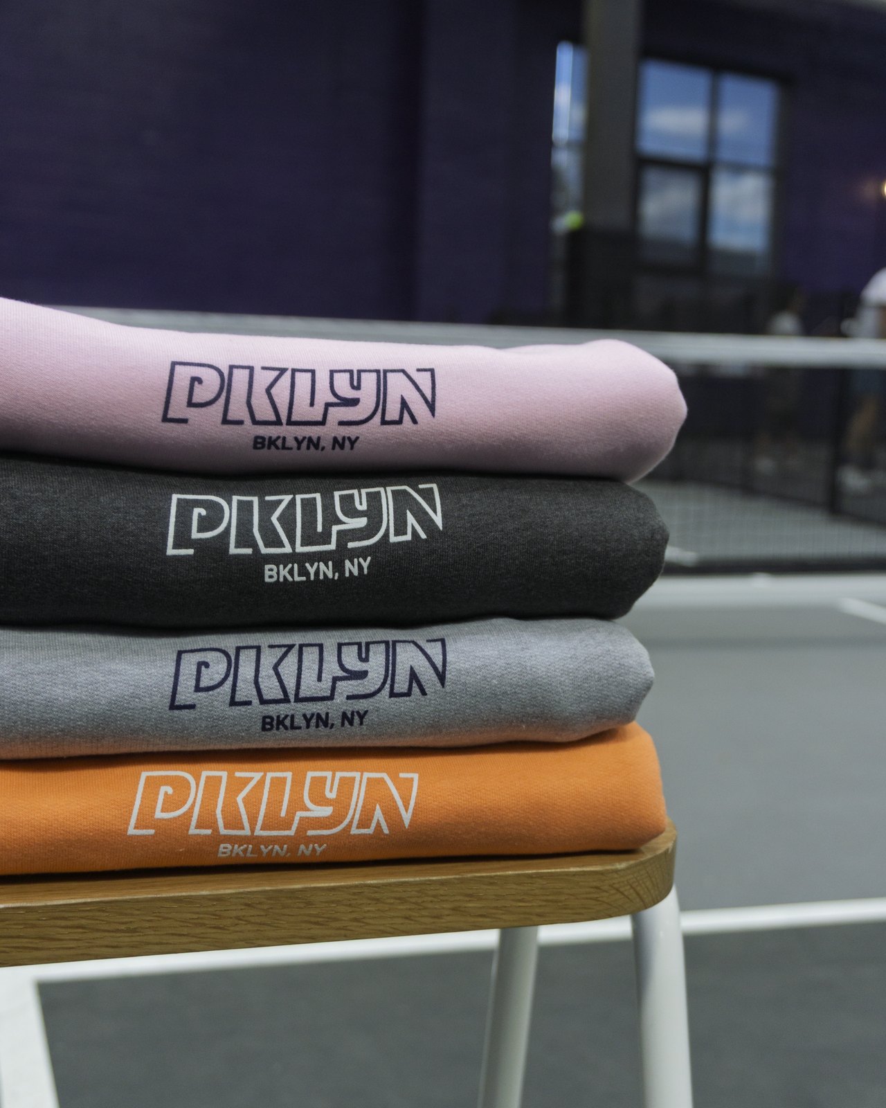Stack of PKLYN sweatshirts in pink, charcoal, grey, and orange